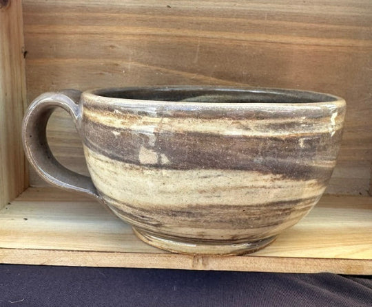 A rustic handmade ceramic mug with a thick handle, featuring a clear glaze over layered clay.
